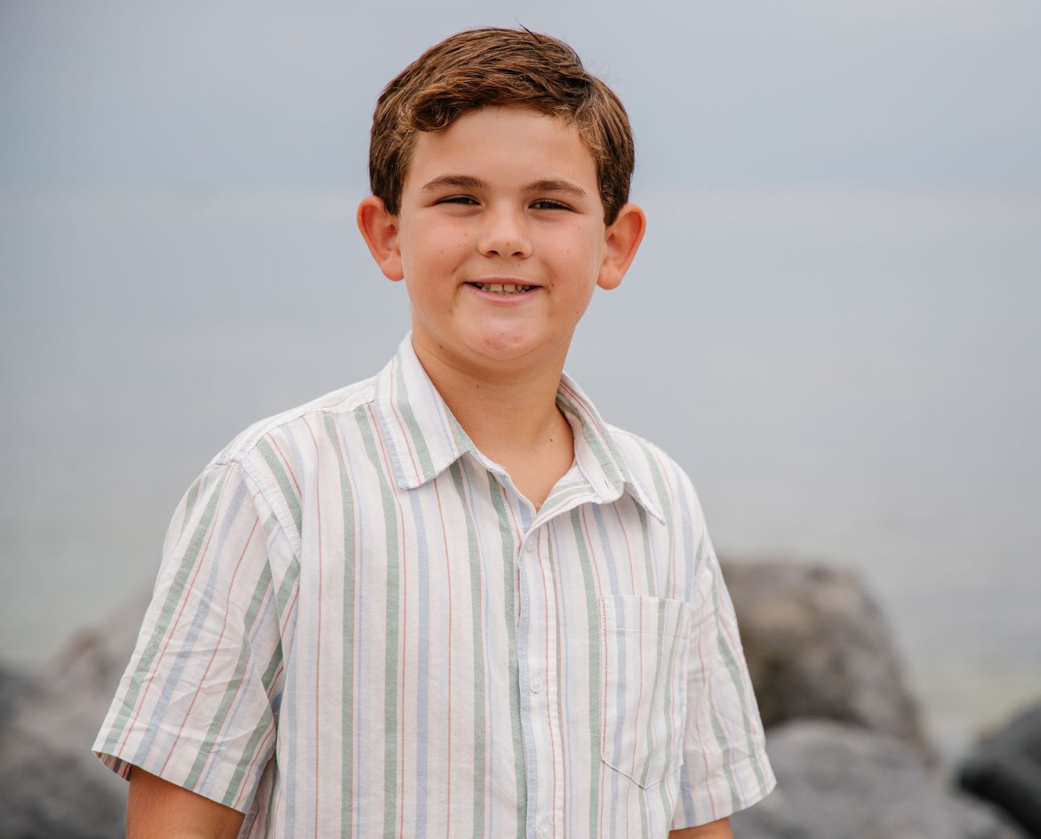 Happy brunette boy in a striped button-up shirt smiling at a rocky beach.