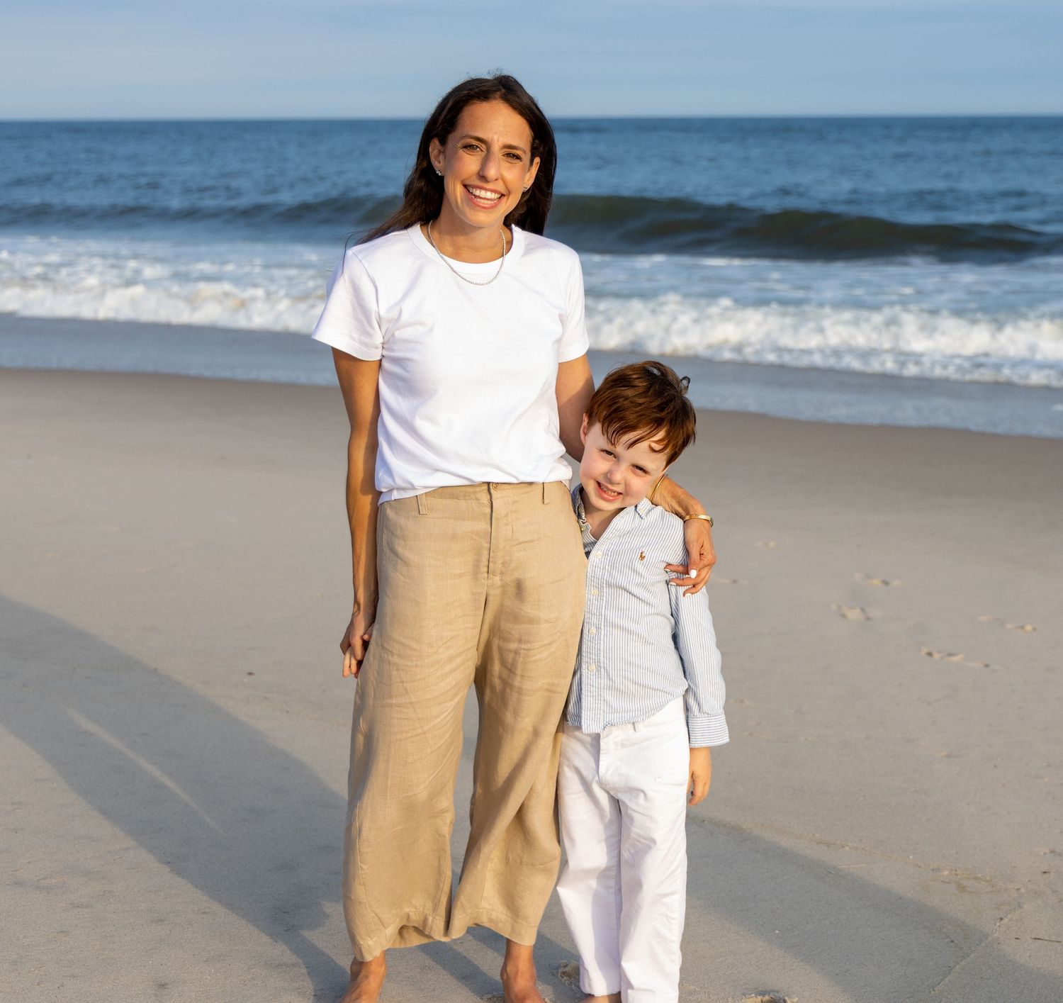 A woman and young boy smile together on a sunny beach, she wears white top and tan pants, he wears light blue shirt. Hamptons photographer