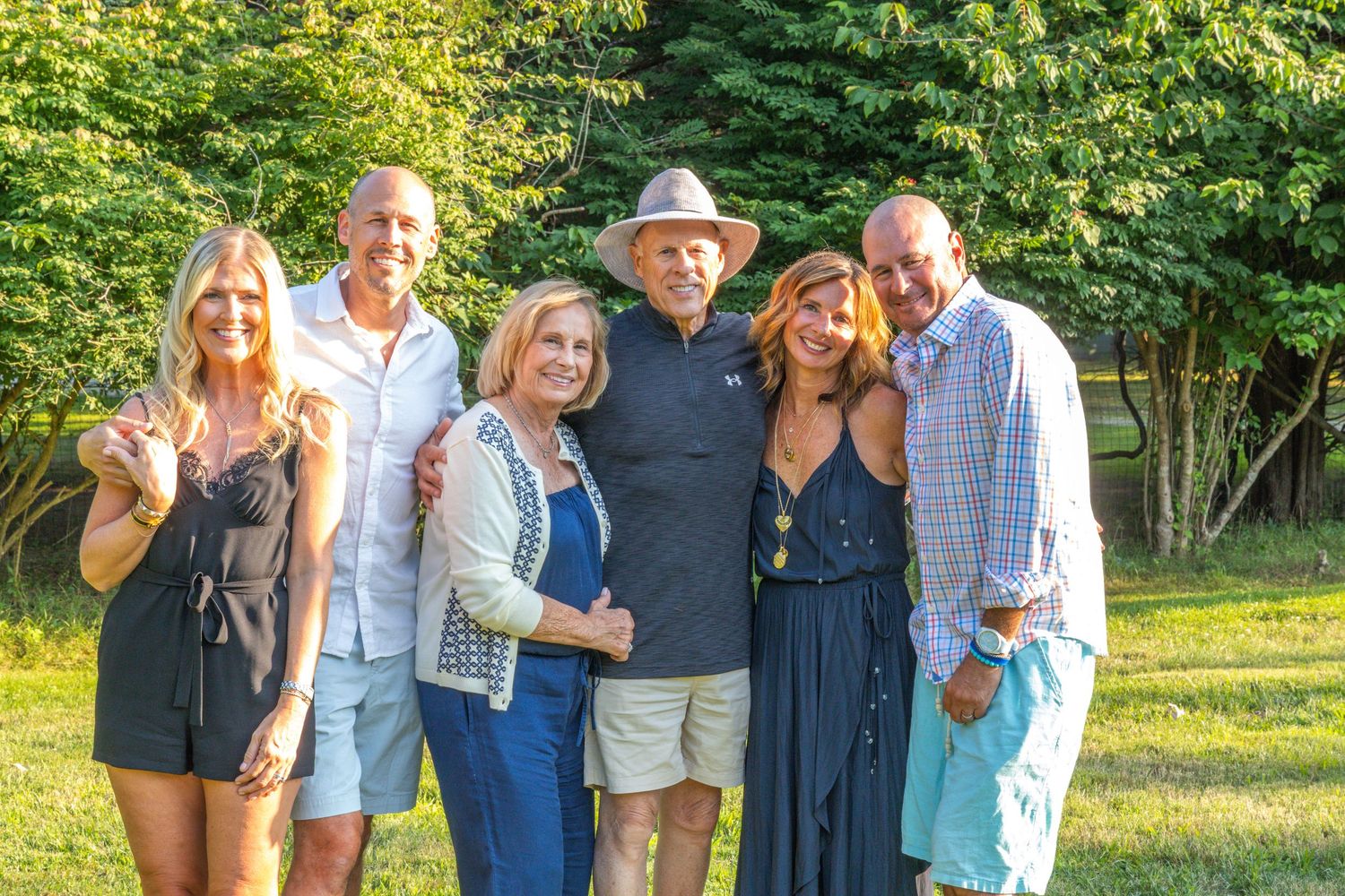 A group of six smiling adults pose together outdoors in a lush green backyard setting.