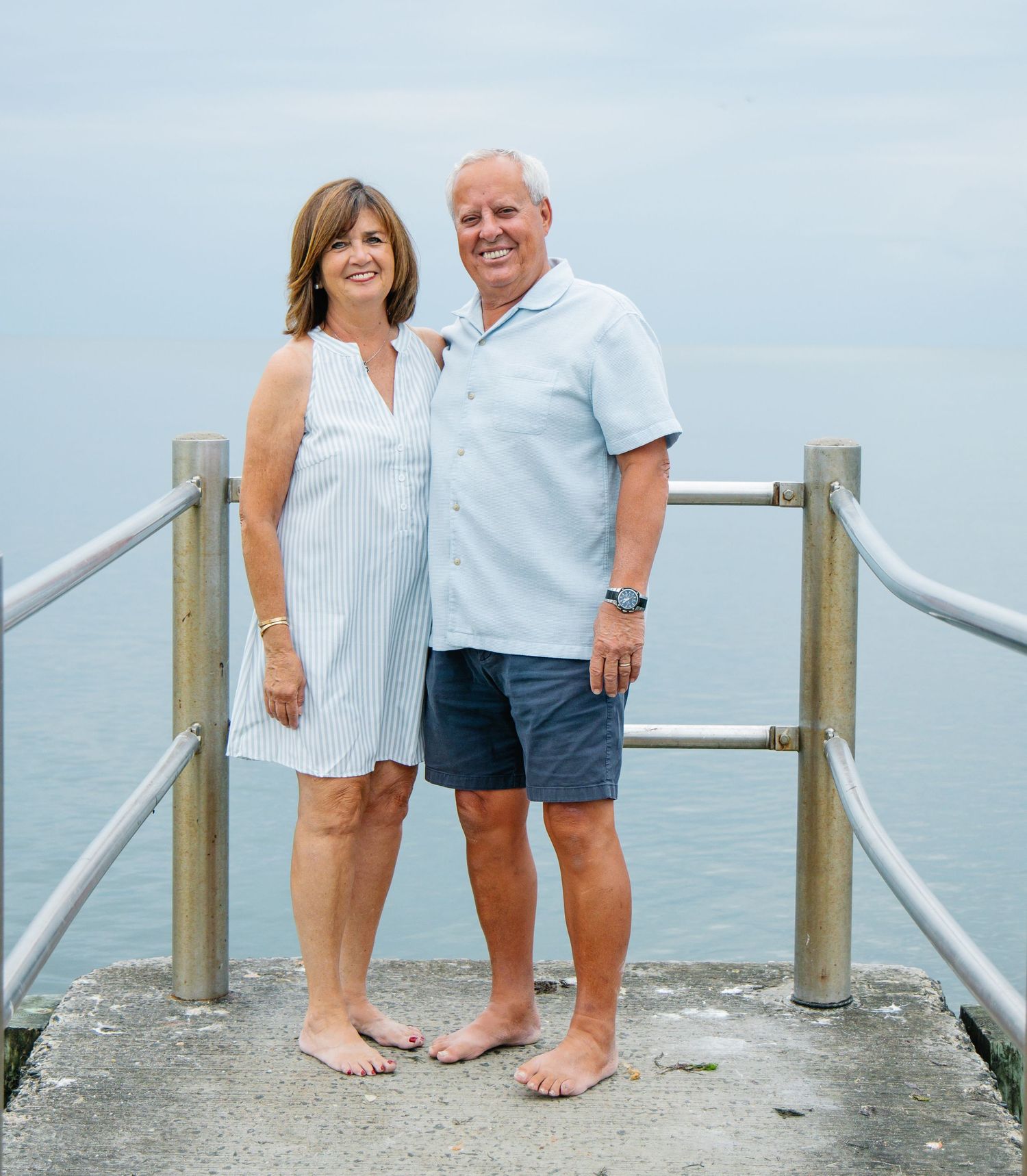 Mature couple standing barefoot on seaside pier, woman in striped dress and man in light blue shirt.