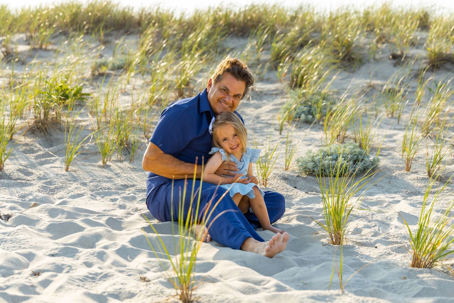 Father and young child sitting together in sandy beach dunes surrounded by sea grass, smiling warmly.