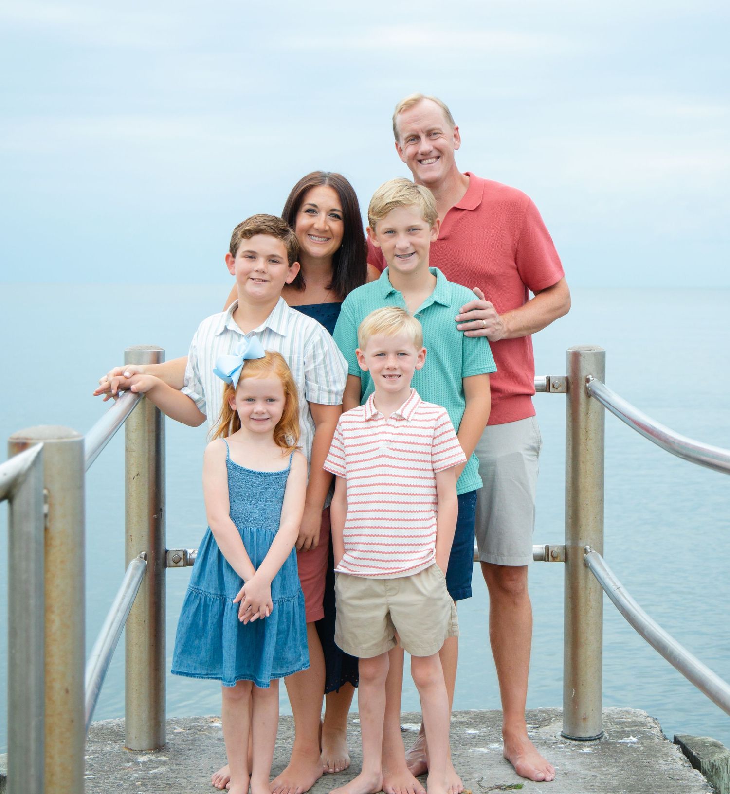Family of six posing on seaside pier with calm blue ocean water in background.