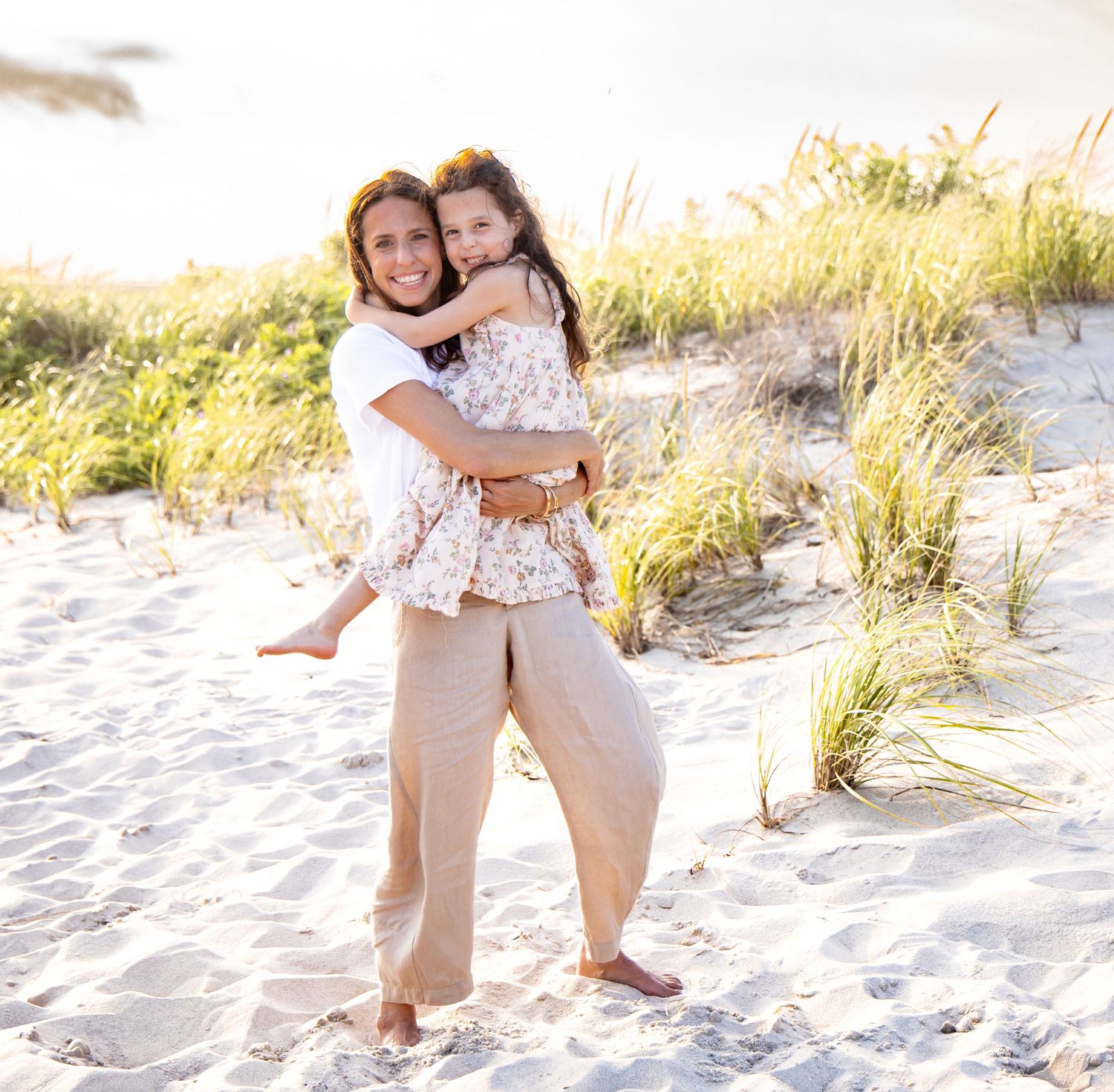 A smiling mother holds her young daughter on a sunny sandy beach with dune grass behind them. Hamptons photographer, family photographer