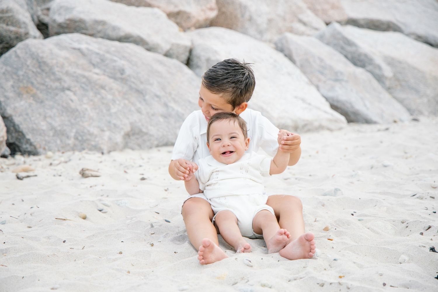 Older sibling sitting behind smiling baby on white sand beach near large rocks.