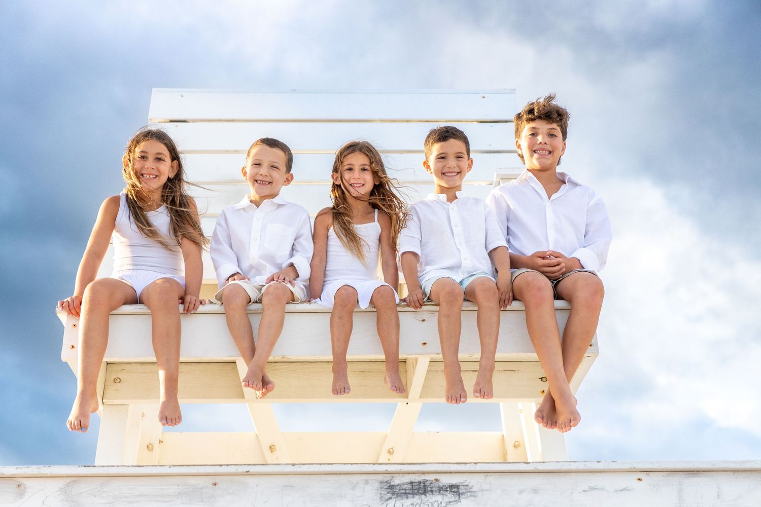 Five happy children sitting on a white lifeguard stand against a cloudy blue sky, wearing white outfits. Hamptons beach. family photographer