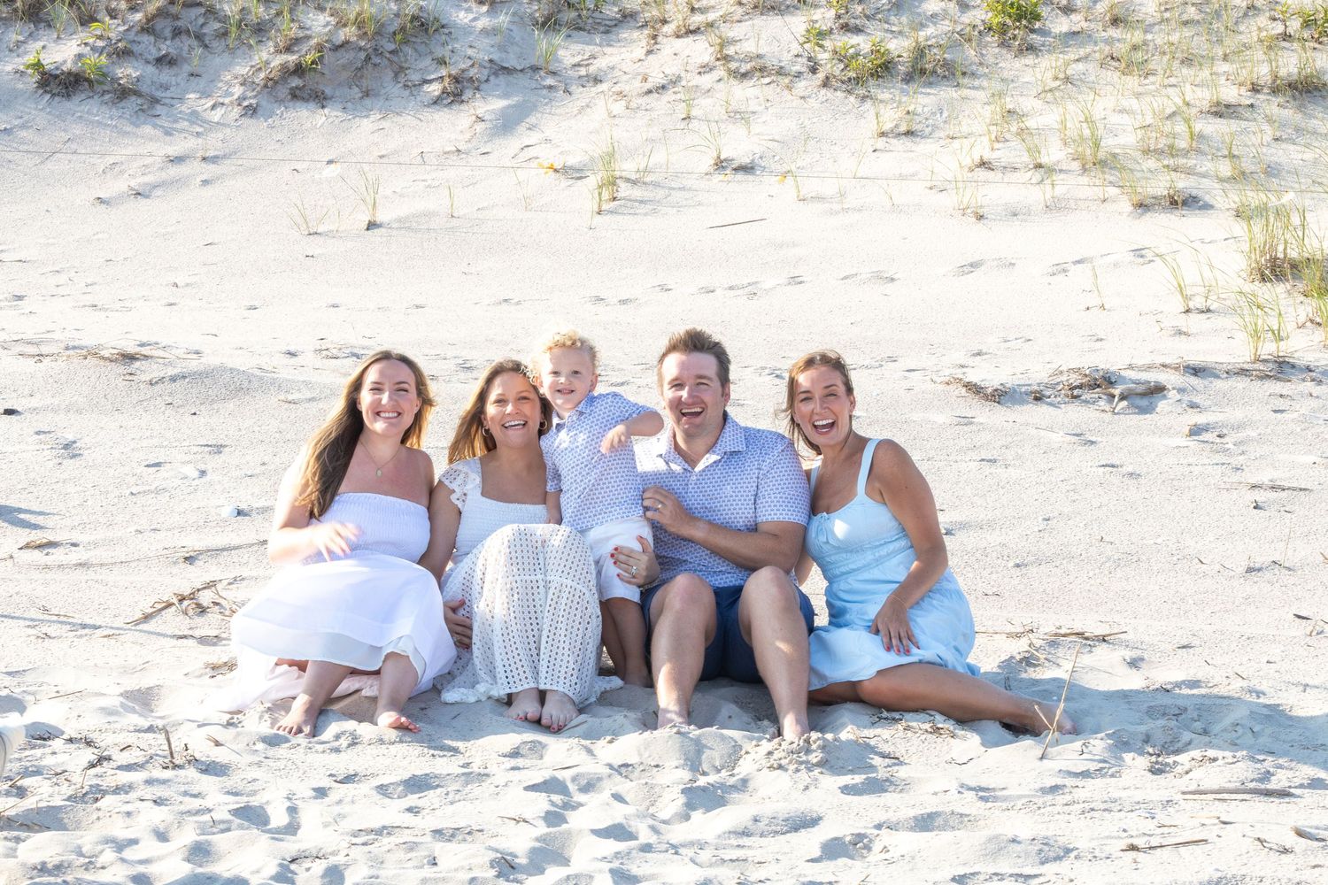 Happy family of five sitting together in beach sand dunes, wearing coordinated white and blue outfits.