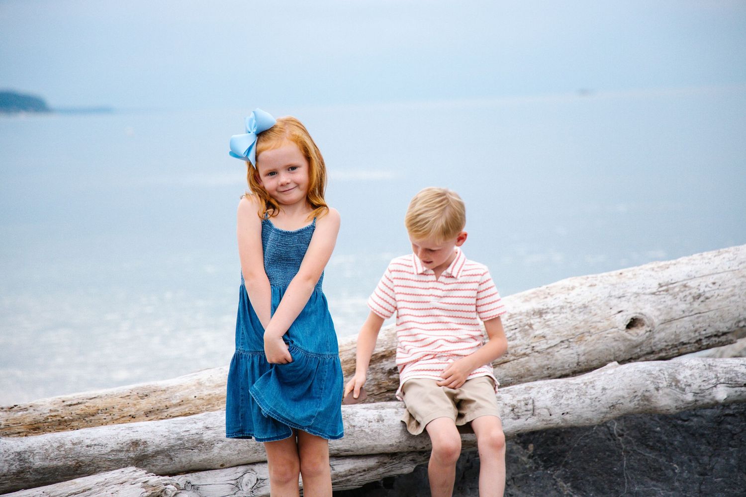 Red-haired girl and blonde boy posing on driftwood log with calm ocean behind them.