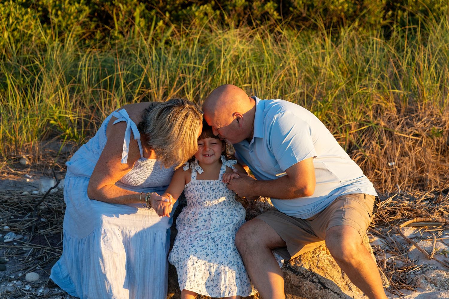 Family of three sharing a tender moment on a sandy beach at golden hour, parents kissing child's cheeks. north fork photographer. family