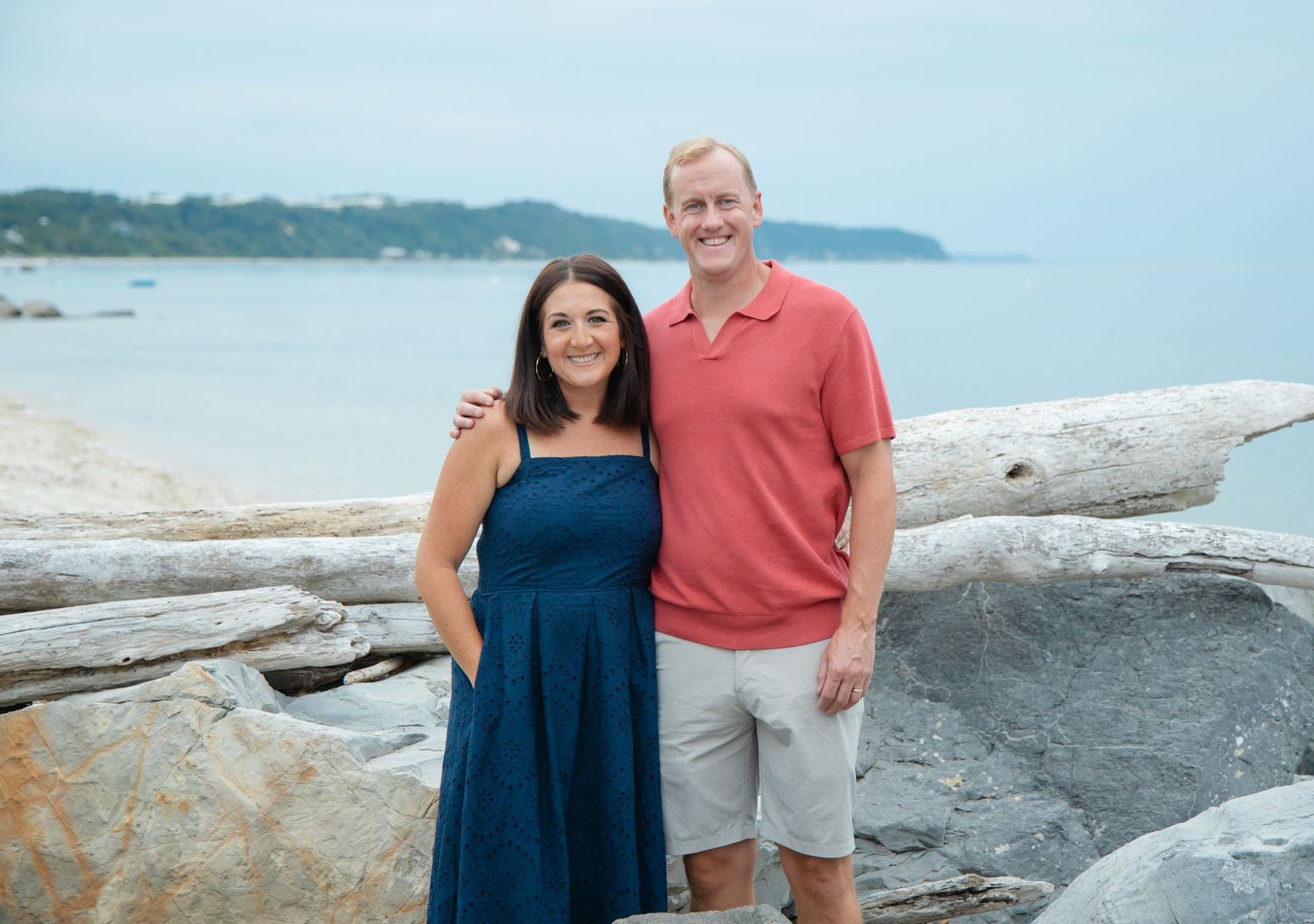 A happy couple embraces on a rocky beach with calm blue ocean waters behind them.