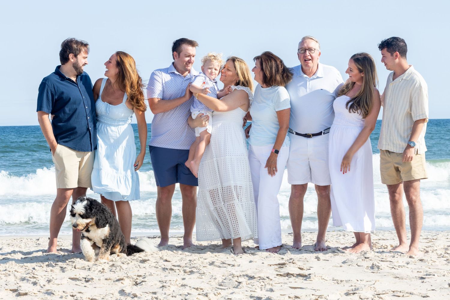 Large family group portrait on beach with dog, wearing white and blue outfits, smiling together by ocean waves.