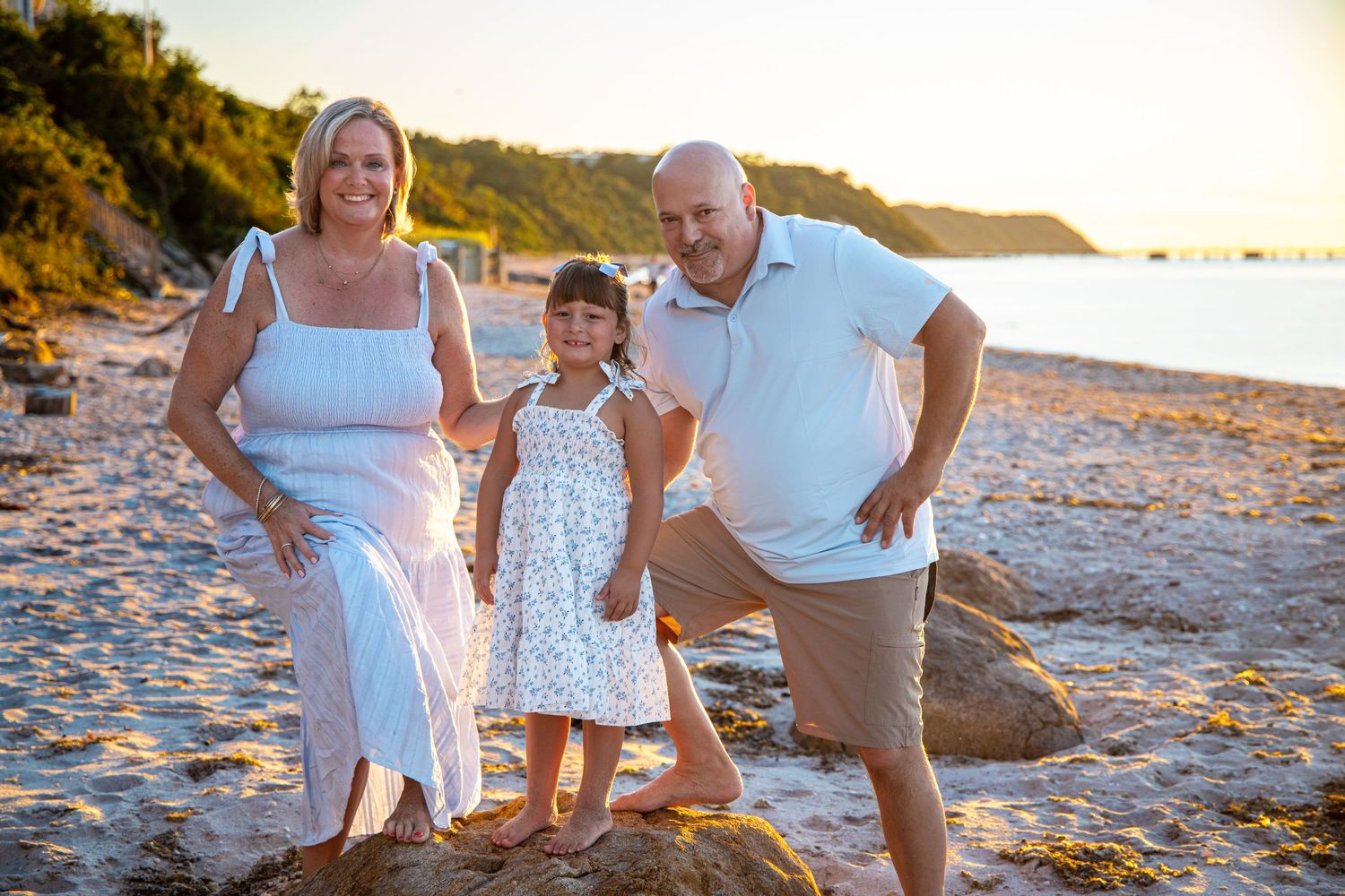 Family of three smiling on a sandy beach at golden hour, woman and girl in white dresses, man in casual attire. north fork photographer.
