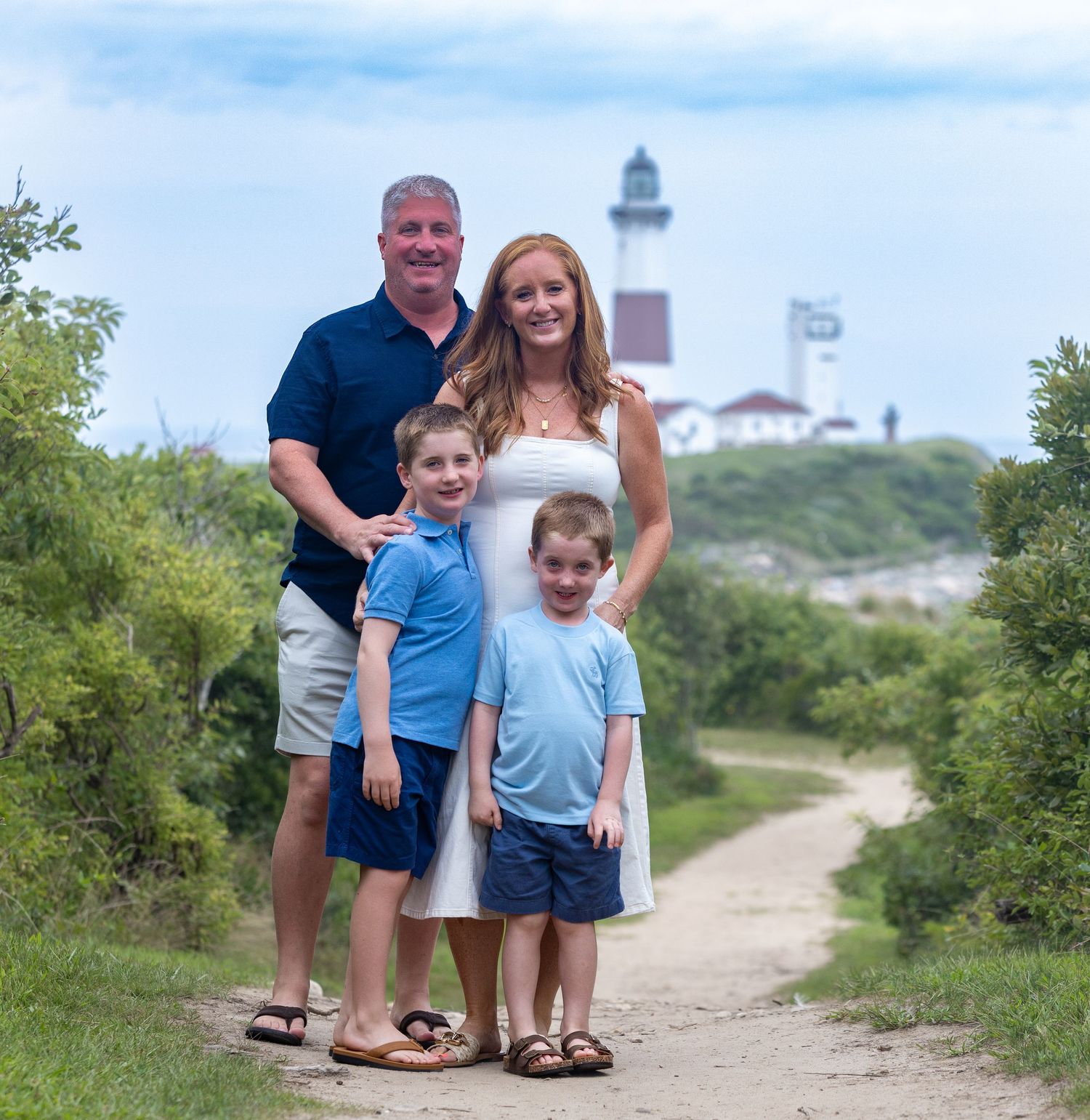 Family of four posing on a scenic path near a lighthouse on a sunny day.