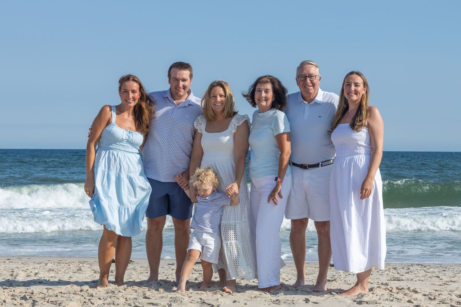 Extended family of seven posing together on sunny beach with ocean waves in background.
