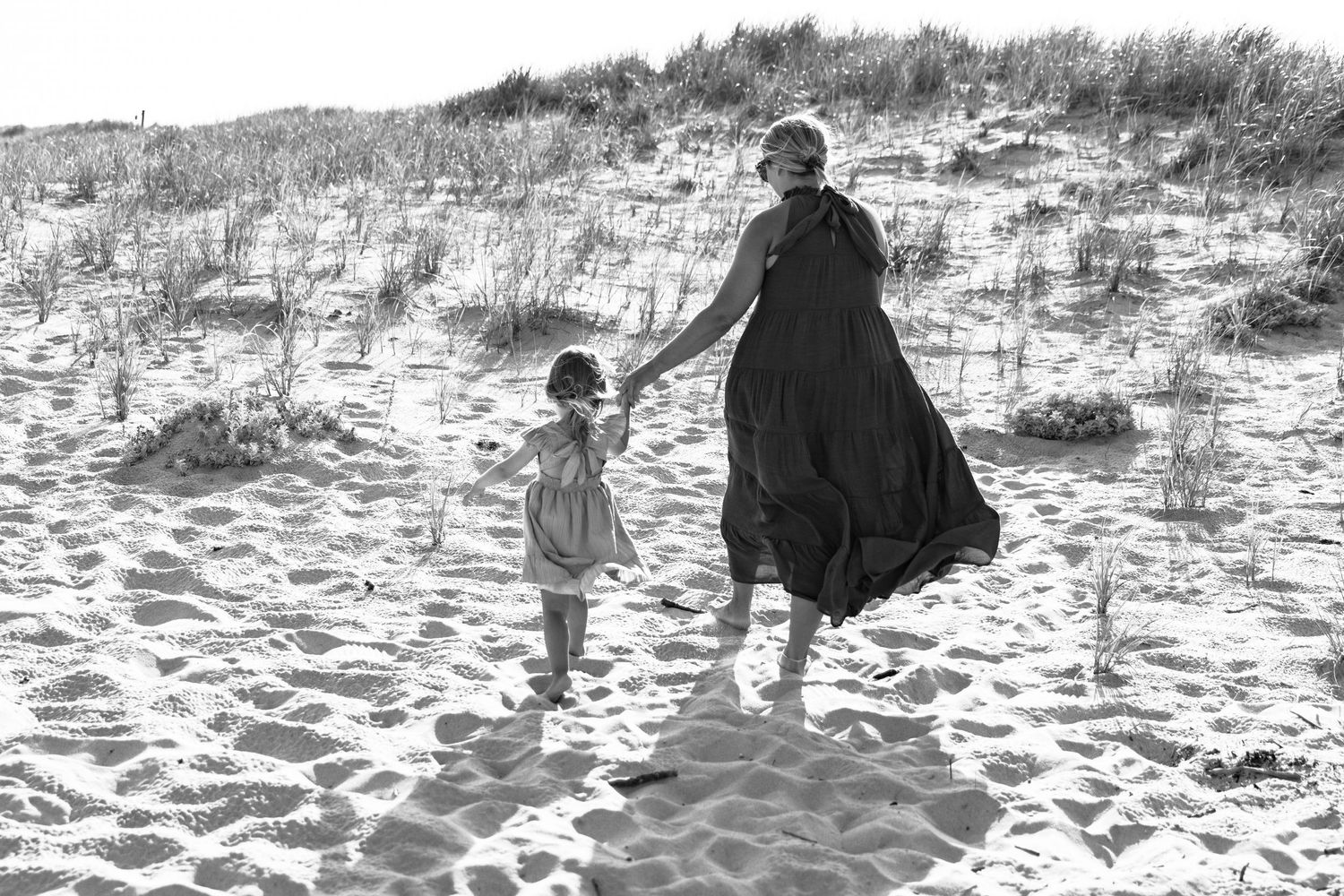 Mother and young daughter walking hand in hand on sandy beach dunes in black and white.