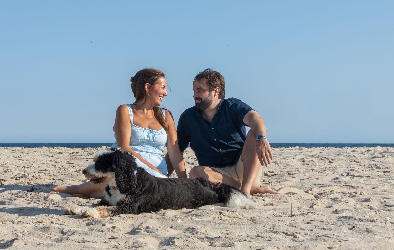 Couple laughing together on beach with their black and white dog between them, sunny clear sky above.