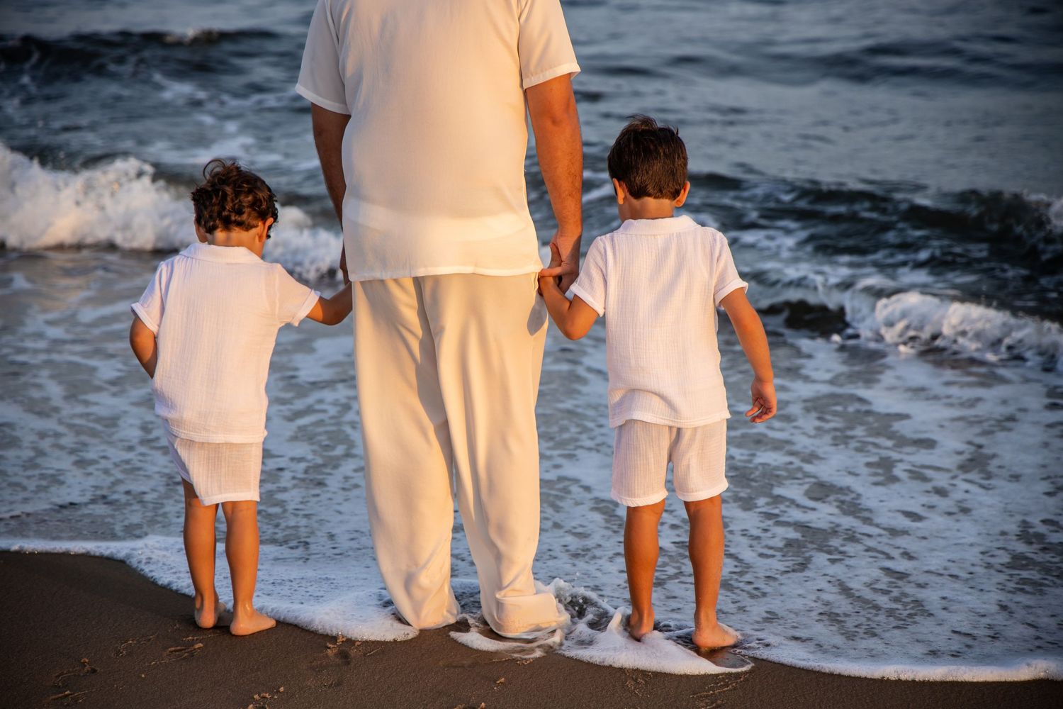 Adult and two children in white clothing holding hands at the beach shoreline with ocean waves. family photographer. Westhampton. Hamptons