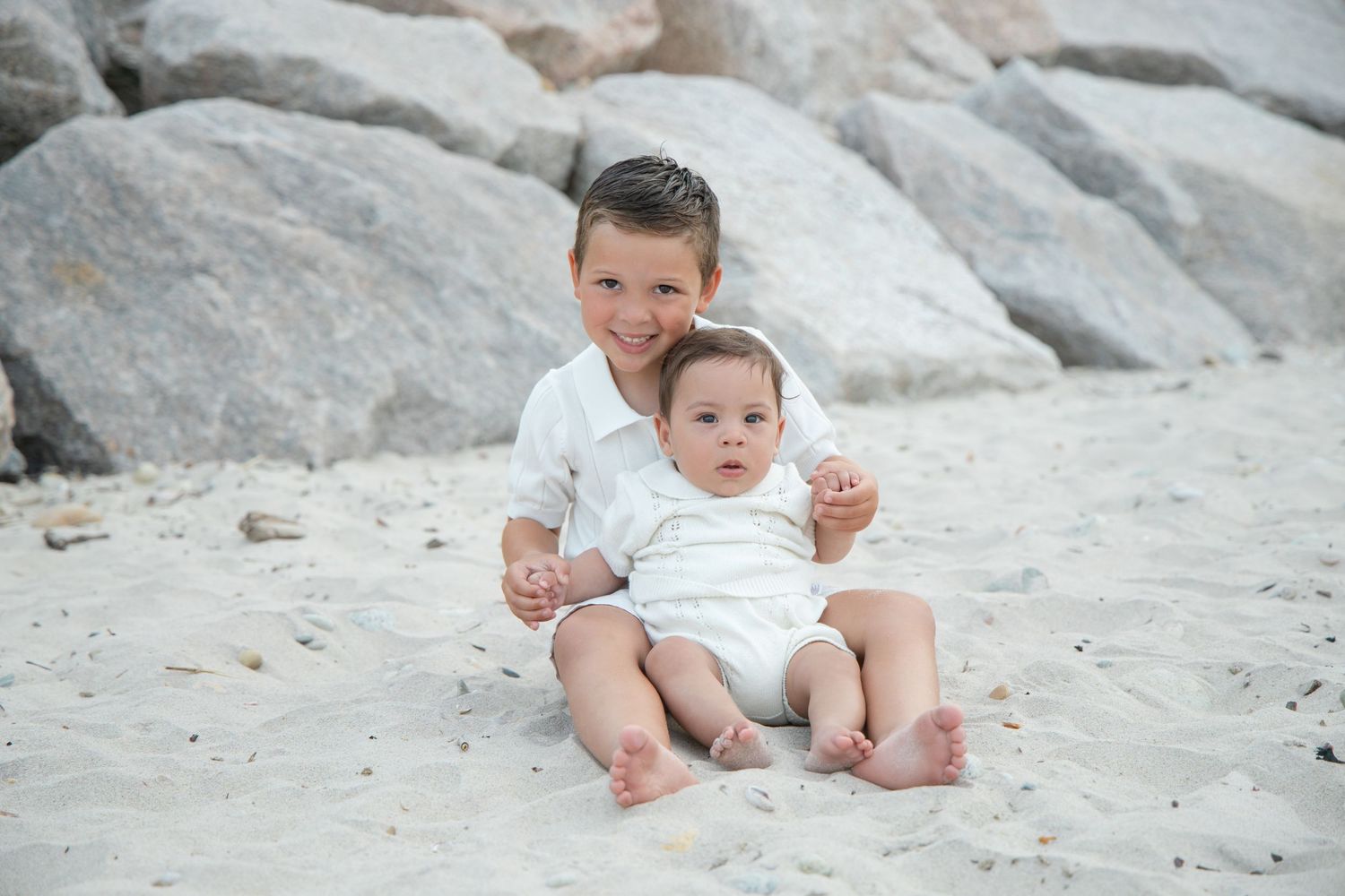 Two brothers in white outfits sitting on sandy beach, older boy hugging younger toddler sibling.