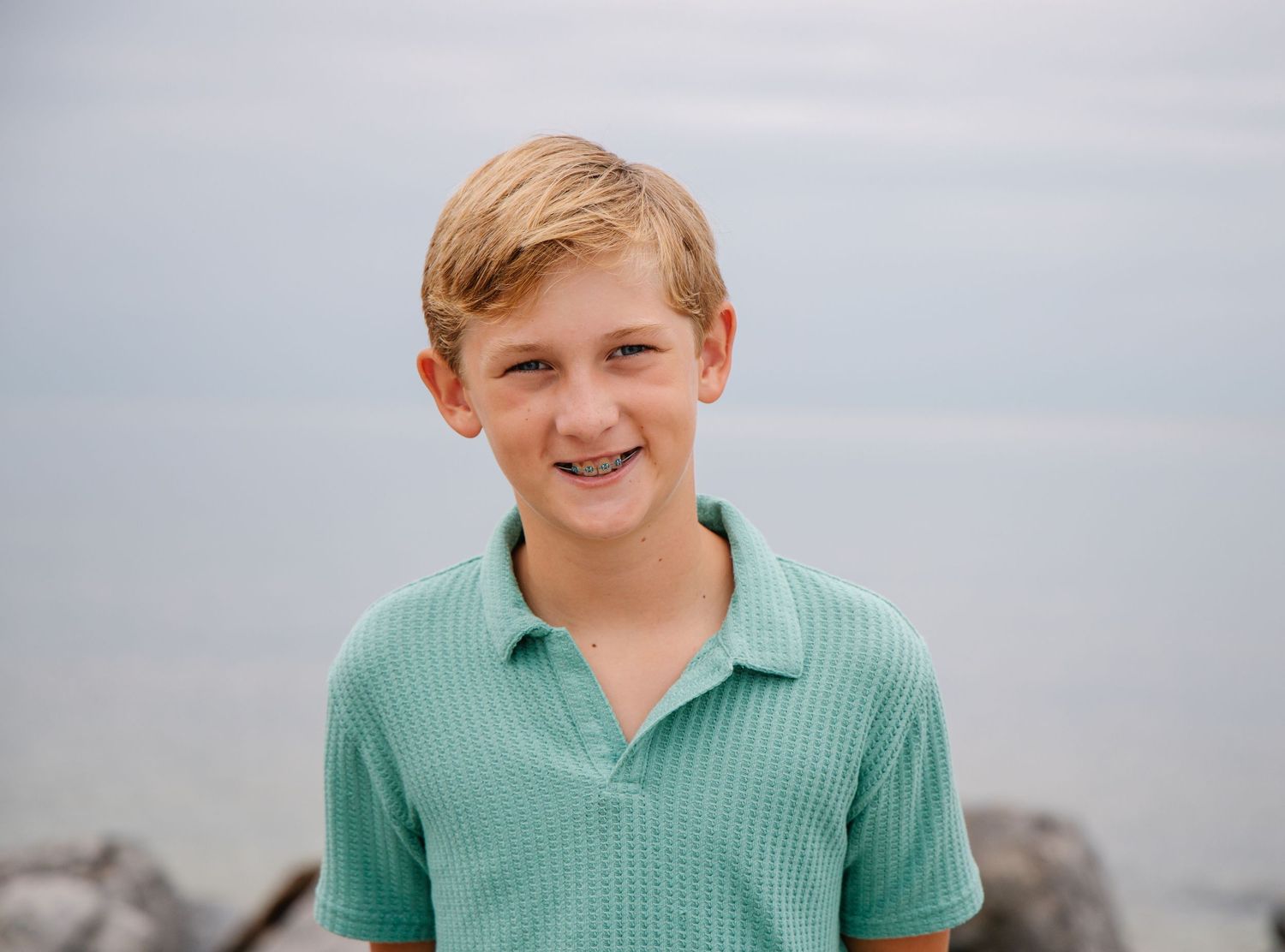 Smiling blond teenage boy wearing a mint green polo shirt at the beach.