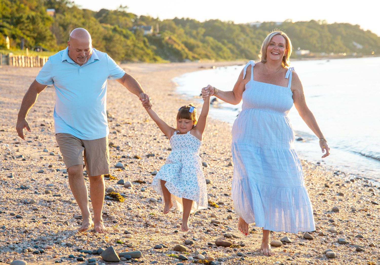 Family of three walking on a sunny beach, parents holding hands with young girl in blue dress.