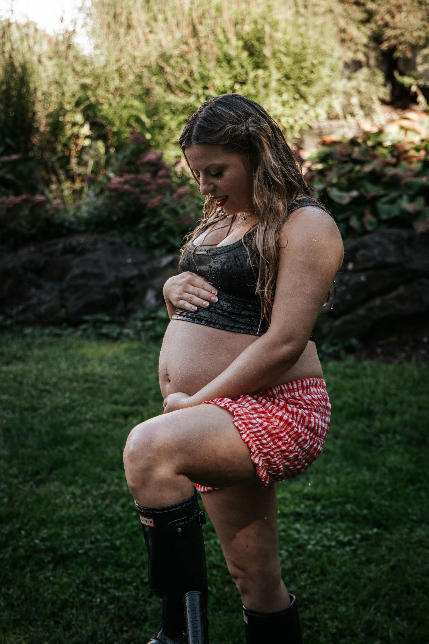 Pregnant woman in black lace bralette and red skirt with black boots cradling baby bump outdoors. Maternity Photographer. Motherhood.