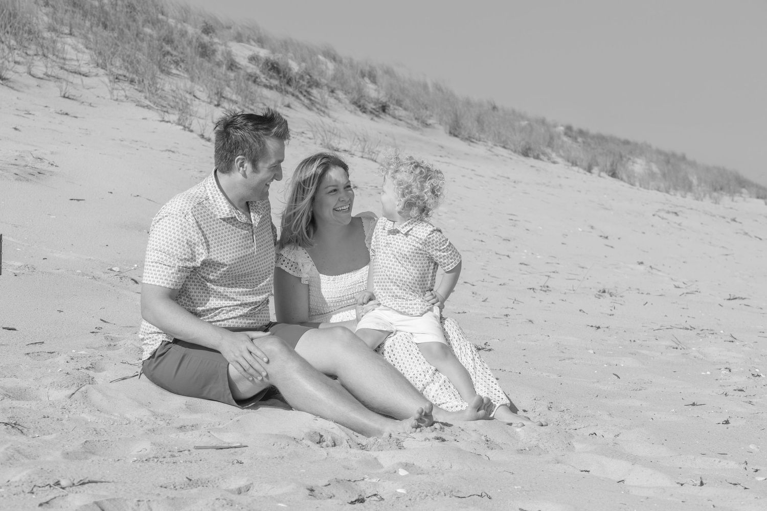 Black and white photo of couple sitting on beach sand with toddler between them.