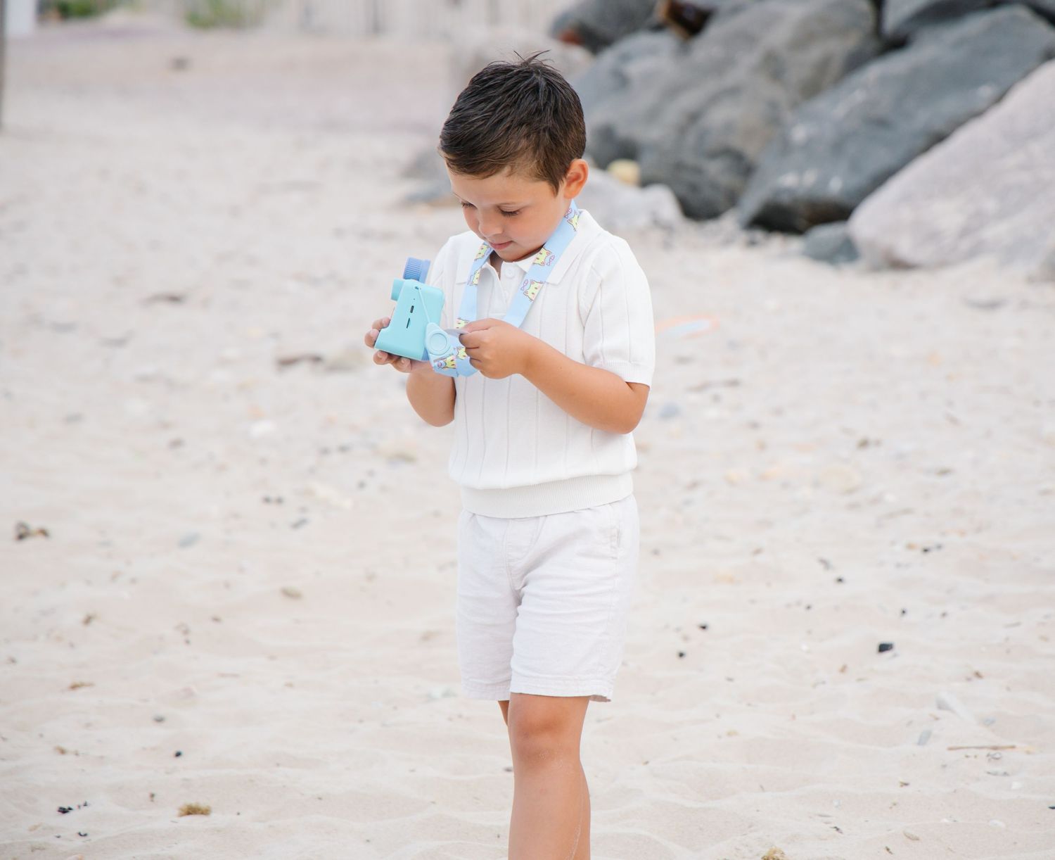 Young boy in white outfit holding a blue toy on a sandy beach.