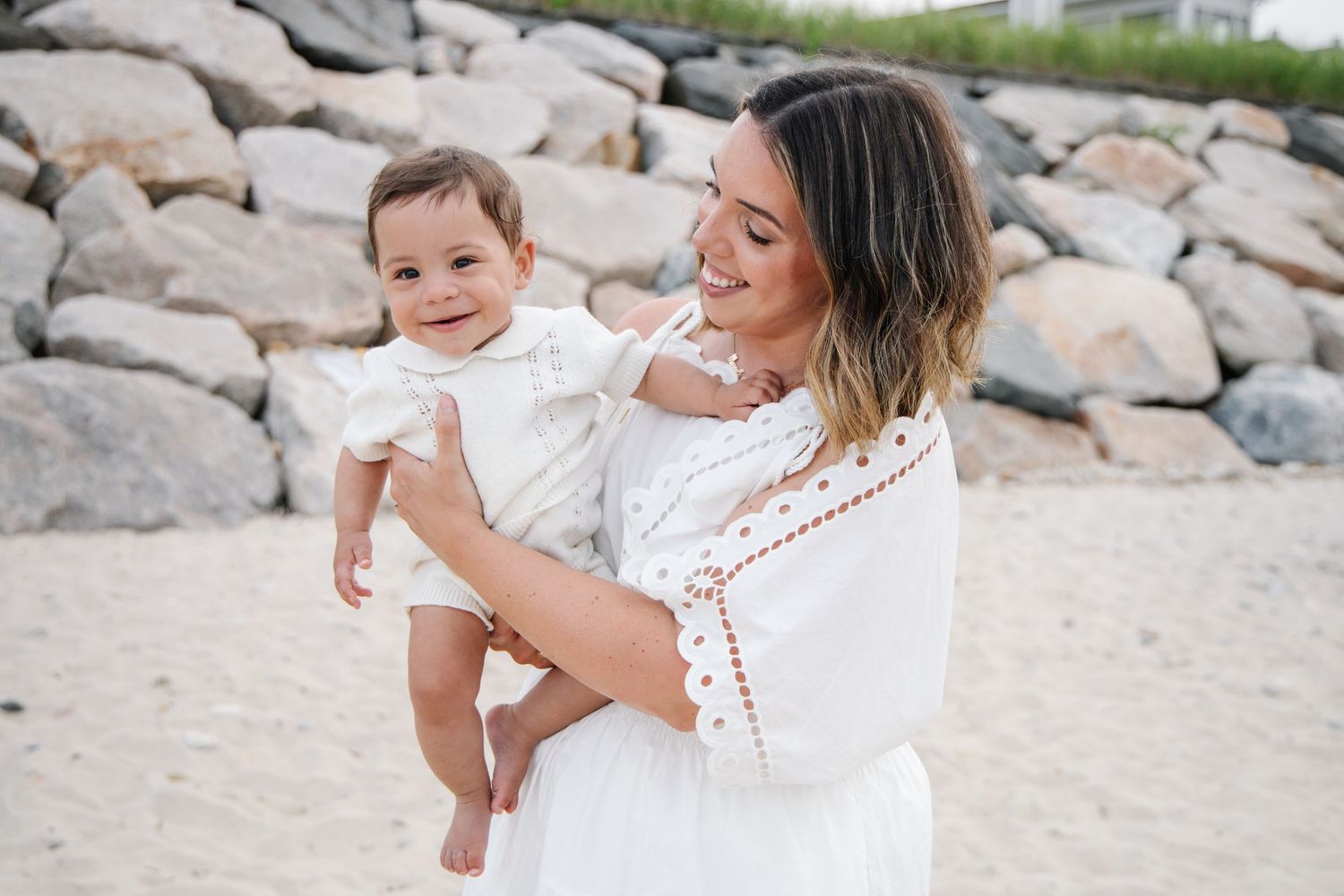 Smiling mother in white lace dress holding laughing baby girl on a sunny sandy beach.