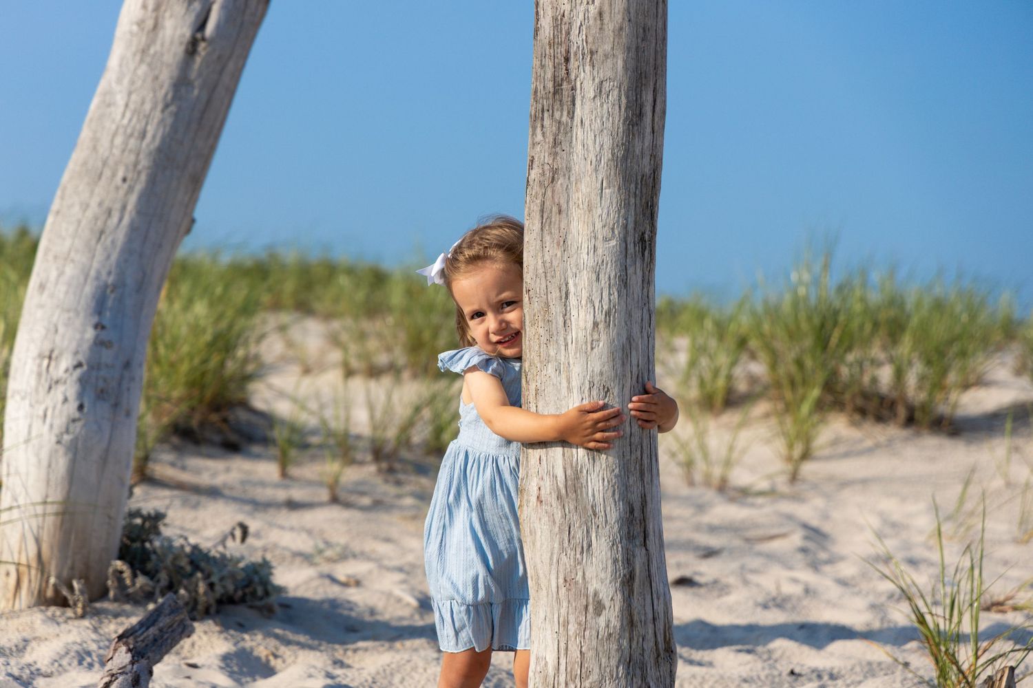 A smiling toddler in a blue romper hugs a driftwood post on a sandy beach dune.