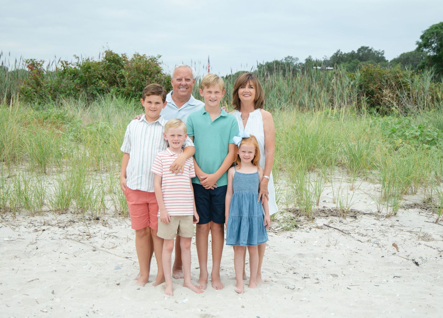 Family of six smiling together on white sand beach with tall grass dunes behind them.