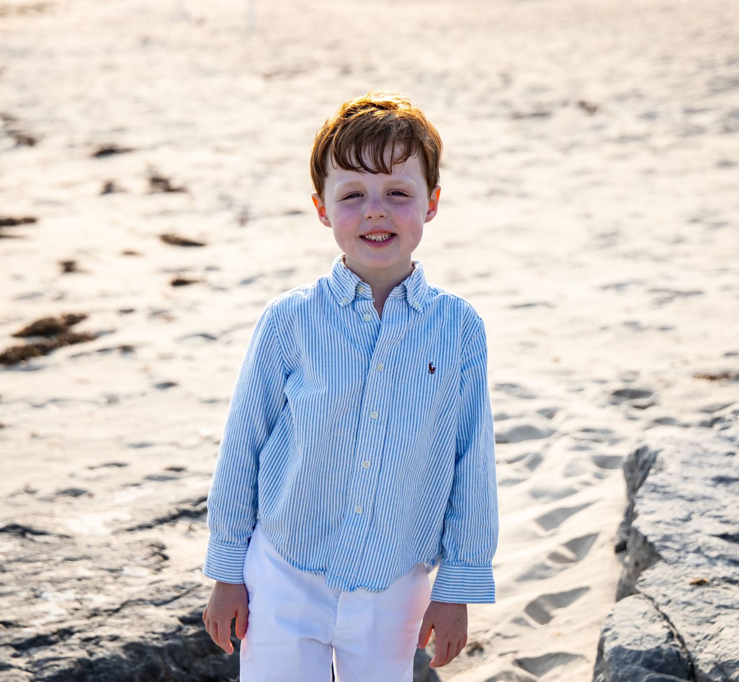 A young boy in a light blue Ralph Lauren shirt and white pants smiling on a sandy beach. family and child photographer. Hamptons beach