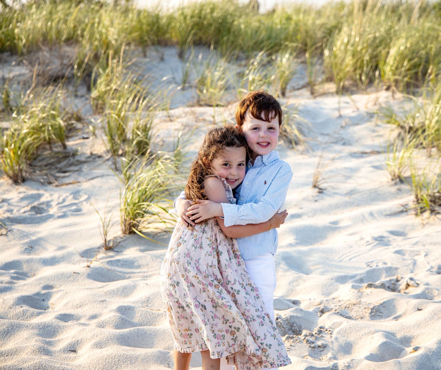 Two children hugging and smiling on a sandy beach with grass dunes in the background on a sunny day. sibling session. Hamptons photographer
