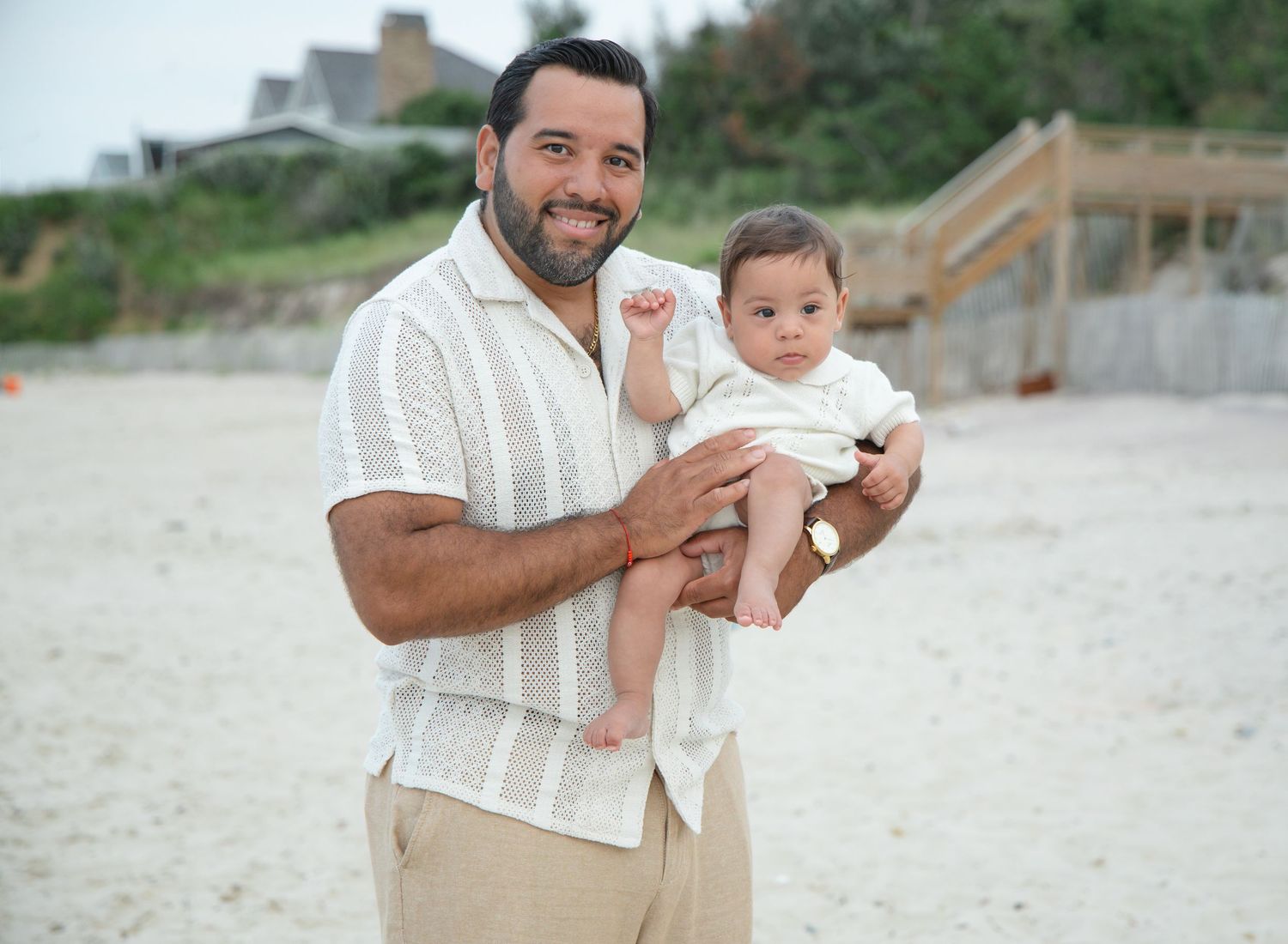 Man holding baby on beach in white outfits, smiling at camera with sandy shore background.