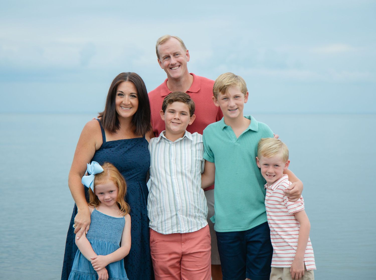 Family of six smiling together at the beach with calm water in the background.