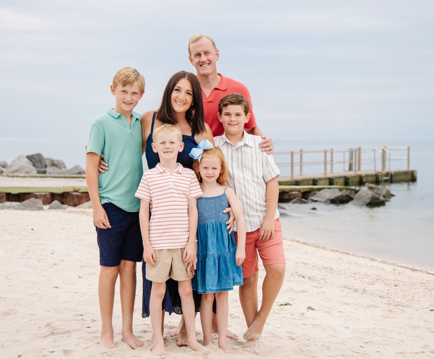 Family of six smiling on sunny beach with rocky jetty and blue water in background.