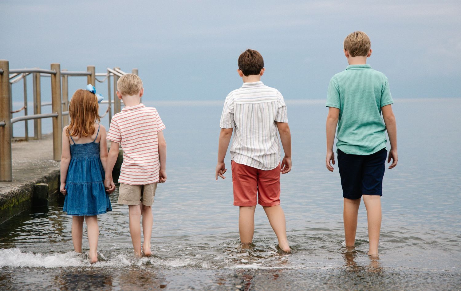 Four children standing at water's edge on a rocky pier, viewed from behind on an overcast day.