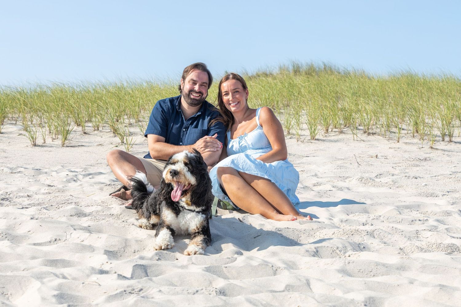 Couple sitting on sandy beach with their black and white dog, smiling at camera with beach grass behind them.