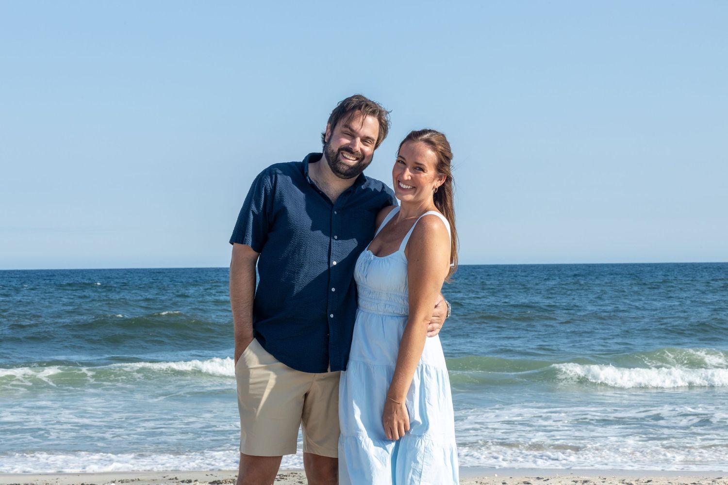 Smiling couple posing on beach, man in navy shirt and woman in light blue dress with ocean waves behind them.