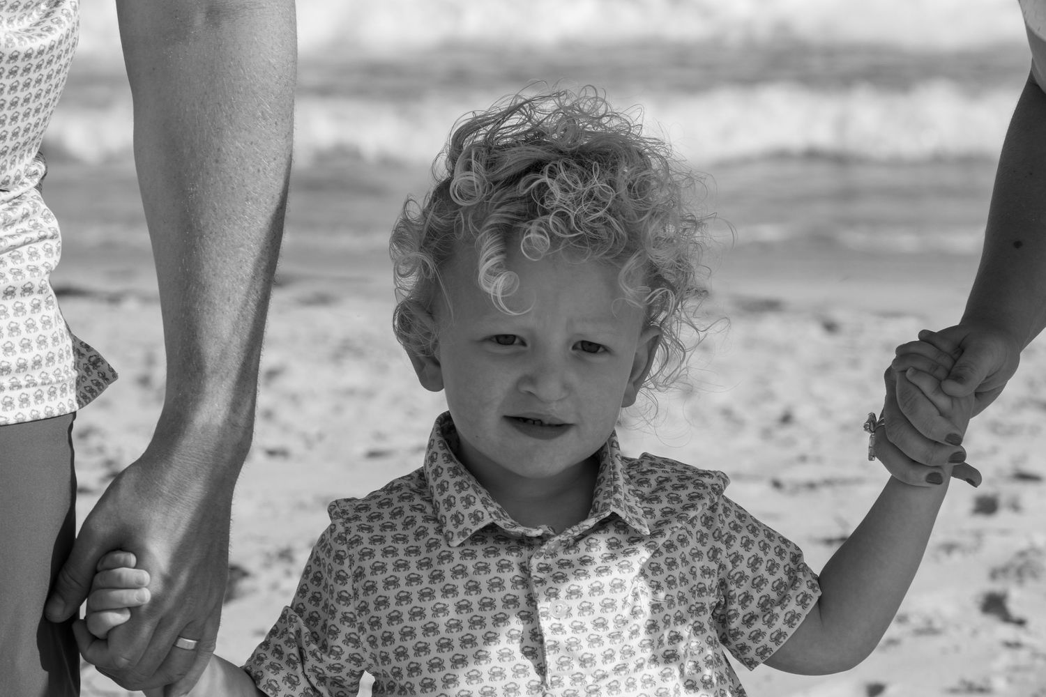 Curly-haired toddler holding parents hands on sandy beach in black and white photo.