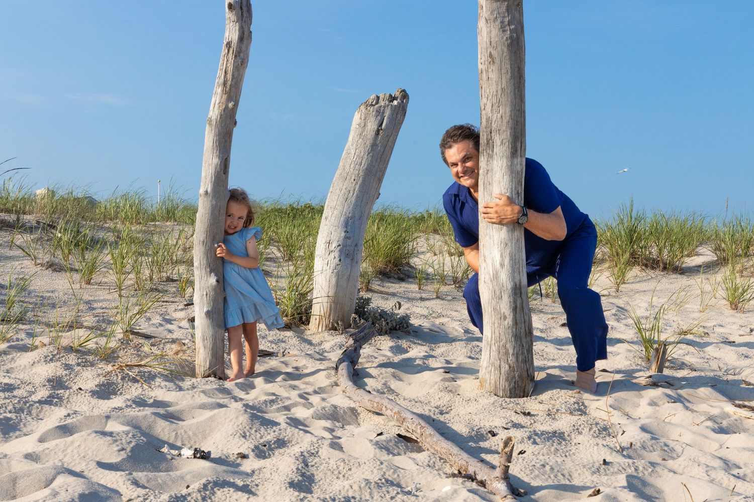 Mother and young daughter playing peek-a-boo around tall driftwood post in sunny beach sand dunes.