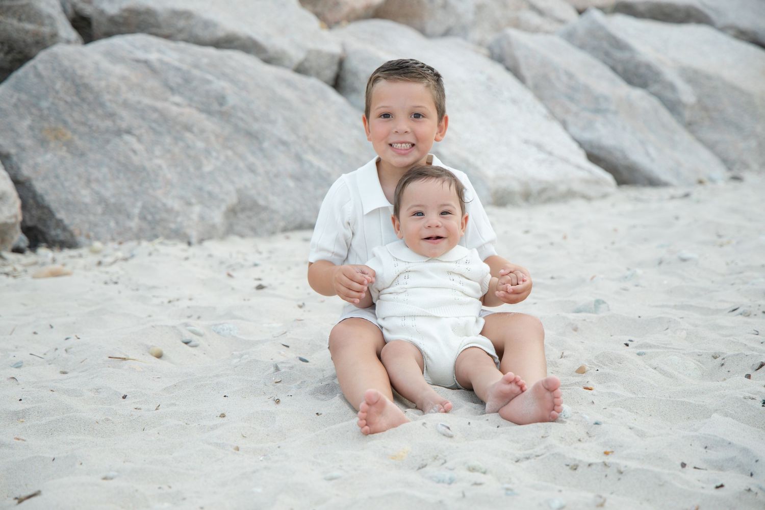 Two young brothers in white outfits smiling together while sitting on sandy beach near rocks.