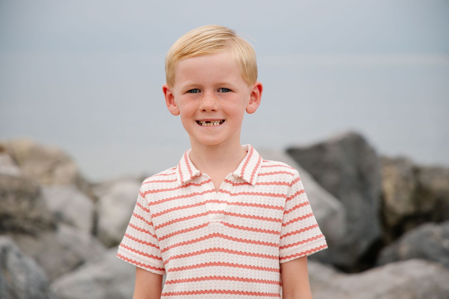 Young blond boy with a gap-toothed smile wearing a striped polo at the beach.