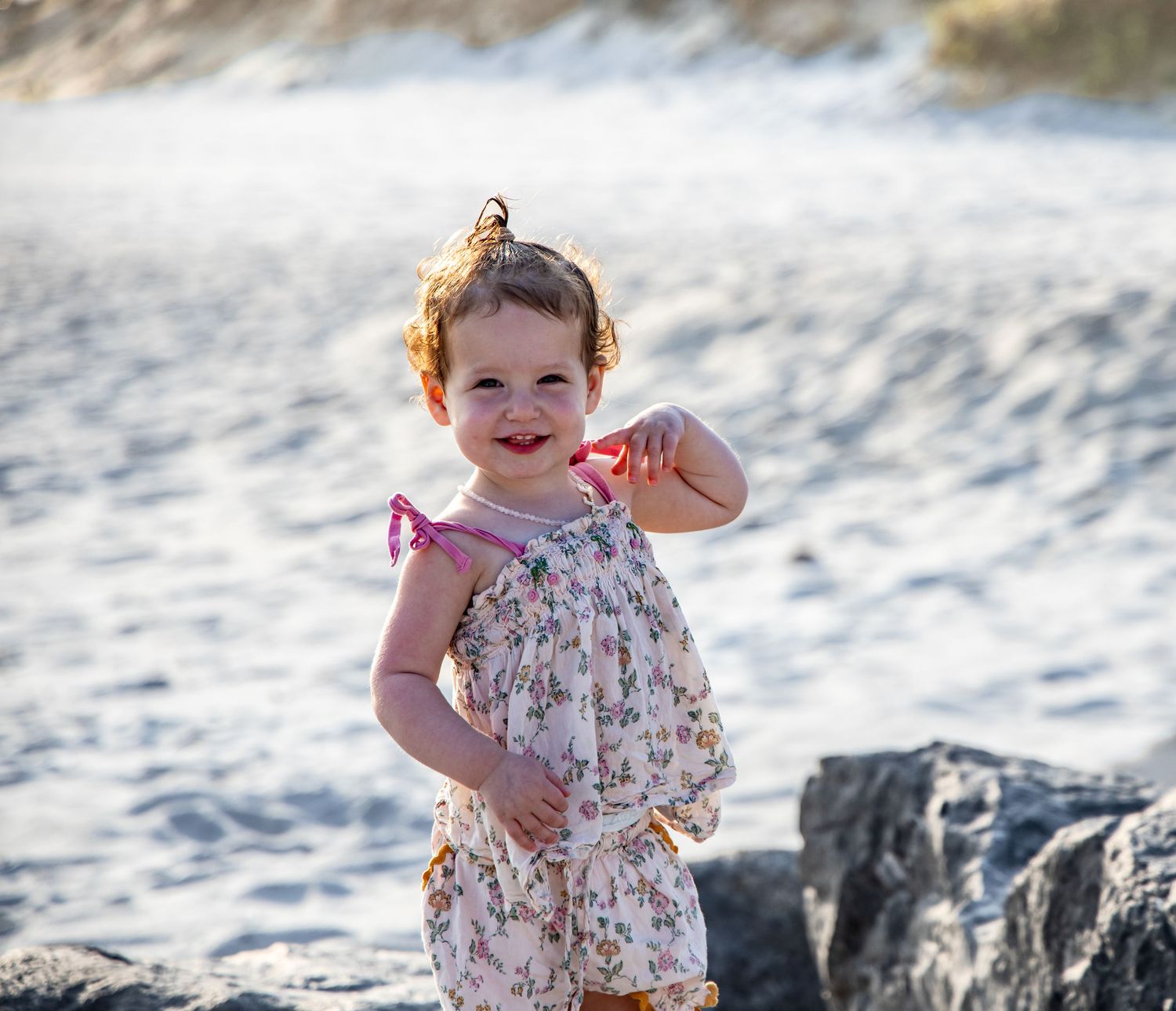 Smiling toddler girl in floral romper posing at sandy beach with rocks in background. Hamptons beach session. family photographer. candid