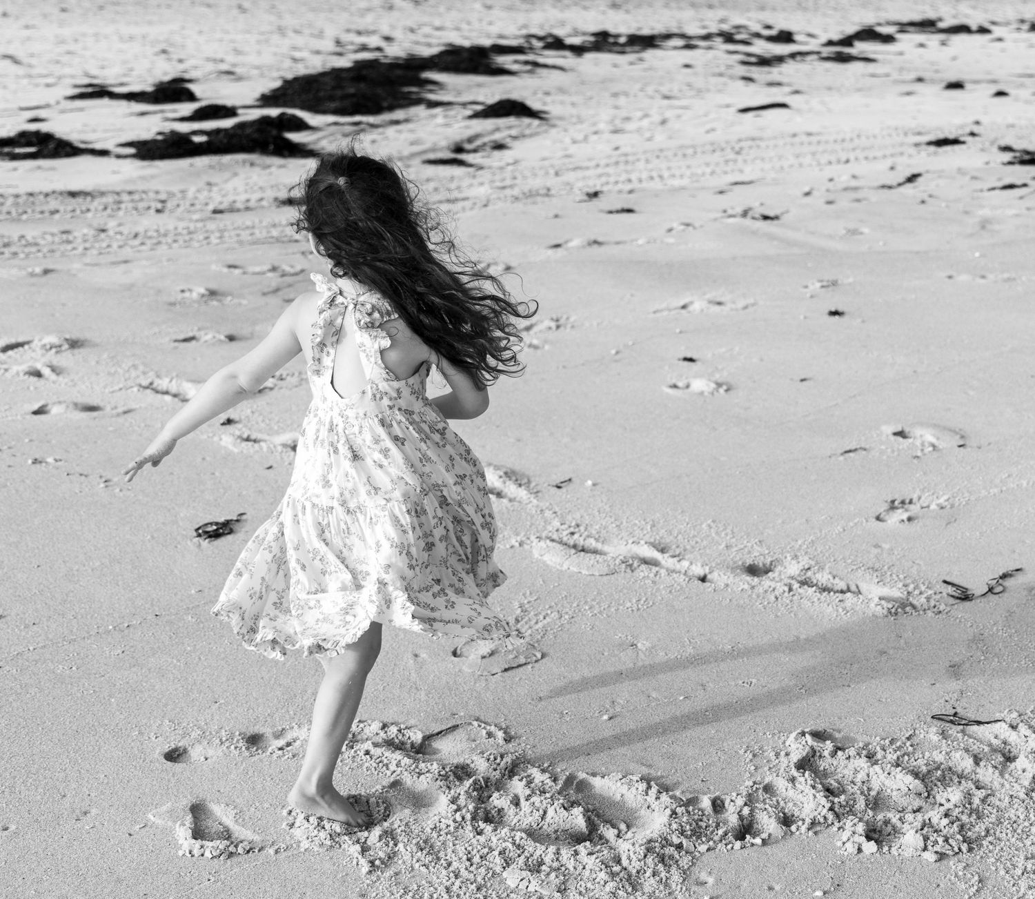 A young girl in a floral dress twirls joyfully on a sandy beach in a black and white photo. child photography, Hamptons photographer. beach