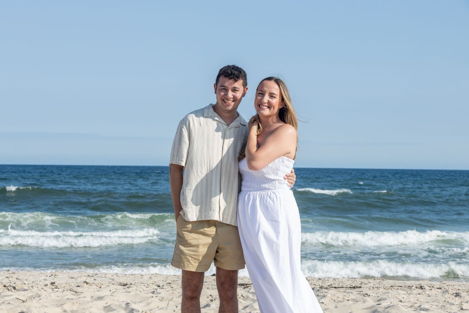 Happy couple embracing on sunny beach, man in striped shirt and woman in white off-shoulder dress, ocean backdrop.