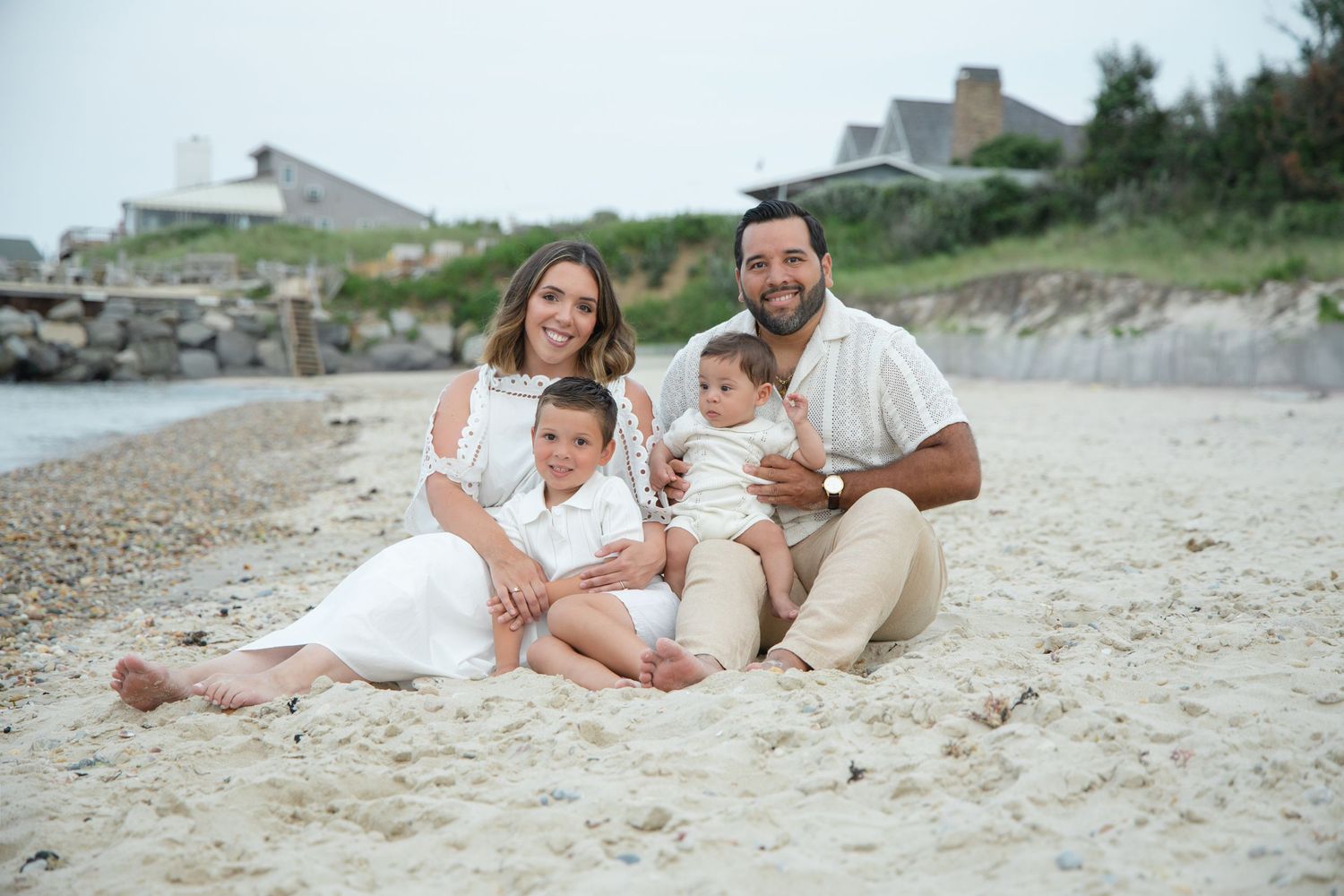 Family of four in white outfits sitting together on sandy beach, smiling at camera.