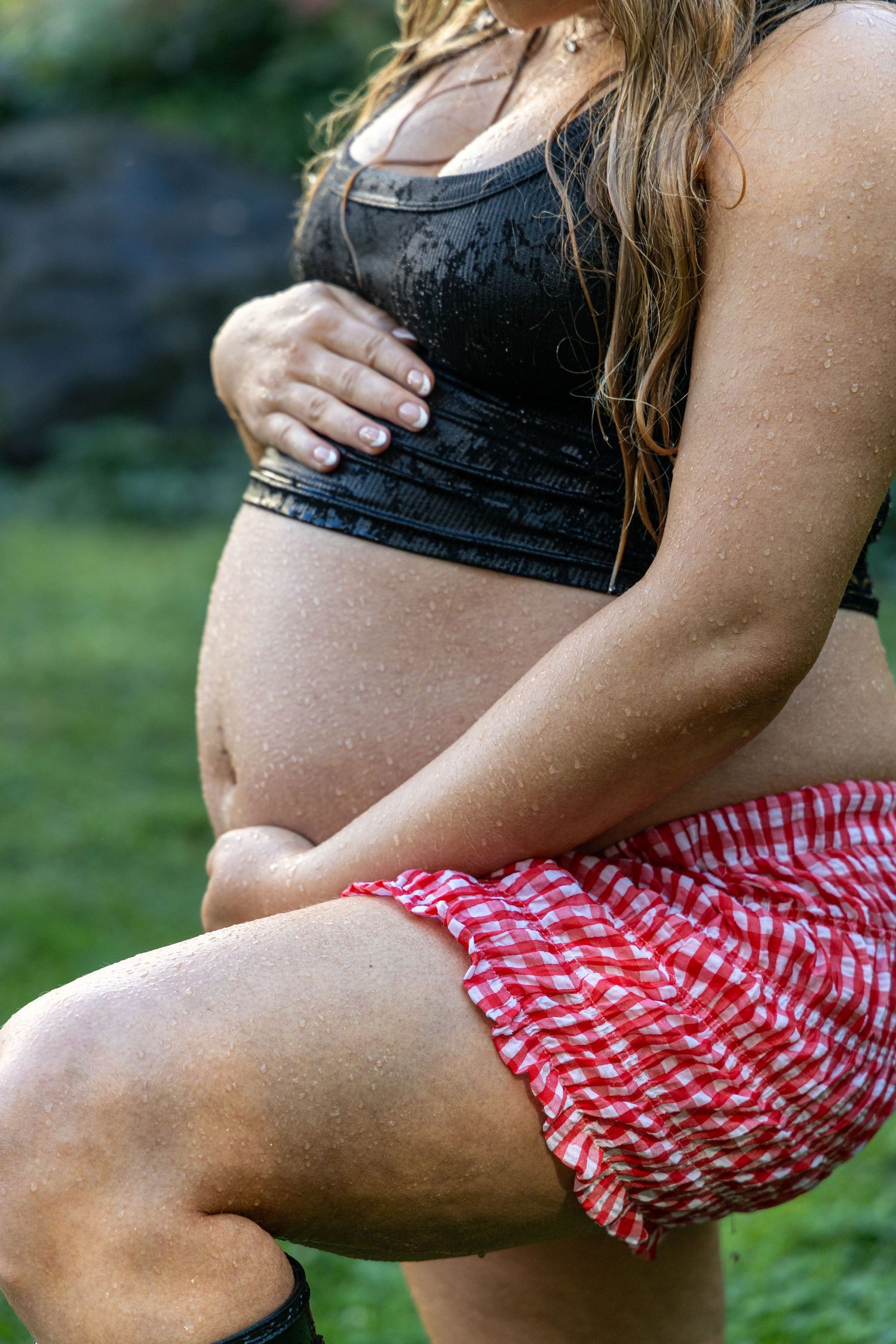 Pregnant woman in black crop top and red gingham skirt cradling her baby bump outdoors in the rain. maternity photographer. motherhood