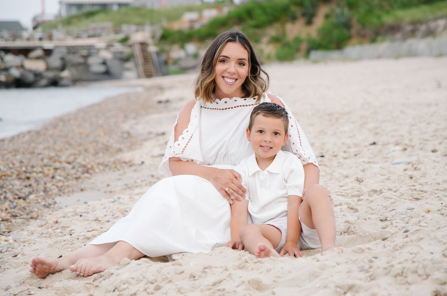 Woman in white dress sitting on beach with young boy in white shirt, smiling together.
