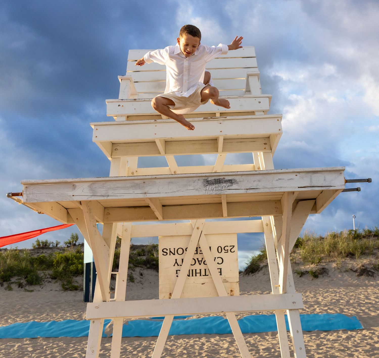 Kid jumping off a white lifeguard stand on a sandy beach under dramatic cloudy skies. Hamptons beach. East Hampton. family photographer.