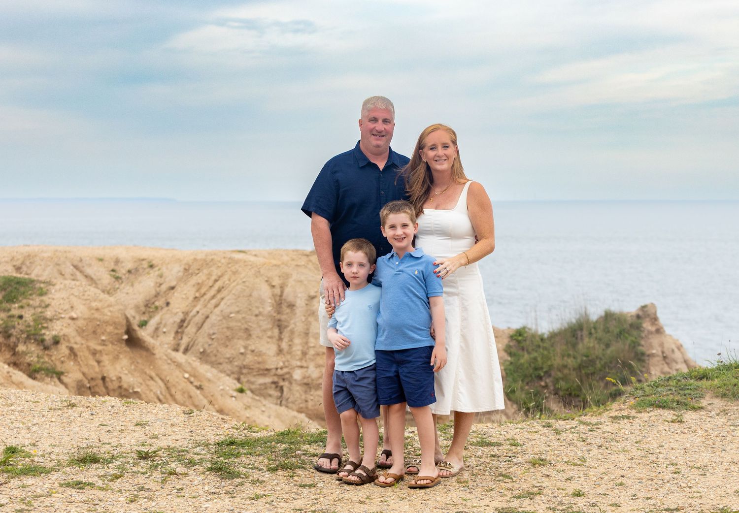 Family of four poses on coastal cliffs with ocean views, parents and two boys smiling in coordinated blue and white outfits.