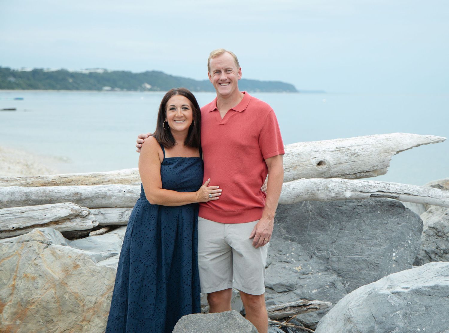 Smiling couple posing on rocky beach with driftwood, ocean and tree-lined shore in background.